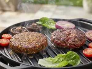 Plant-based burger patty sizzling on grill next to beef patty, with fresh vegetables and steam rising in soft daylight.