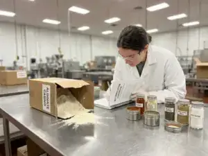 Open alginate package spilling on factory table, manufacturer in lab coat reading guidebook, surrounded by spices under soft lighting.